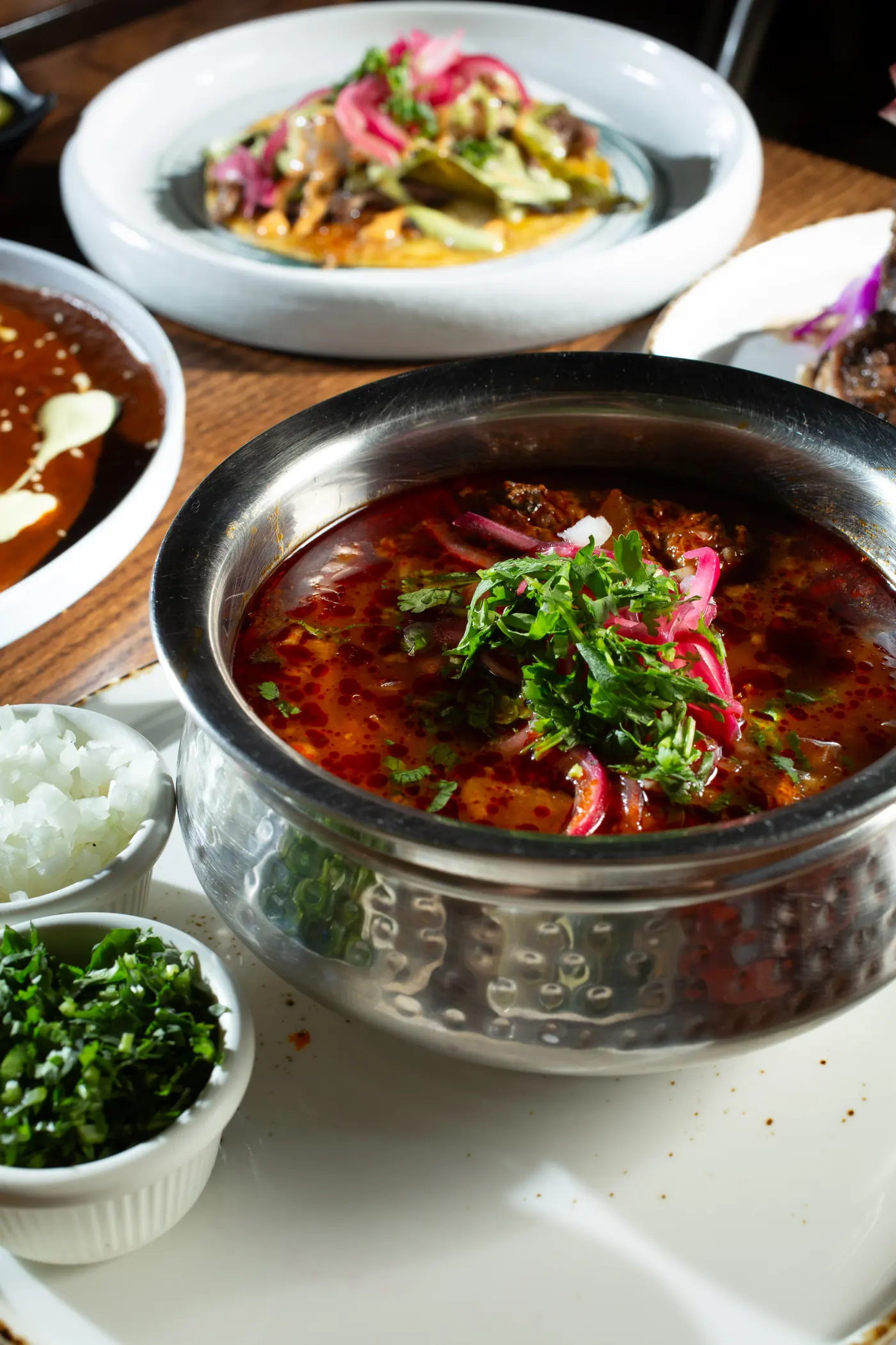 A bowl of birria consommé topped with cilantro and pickled red onion, with chopped onion and cilantro on the side